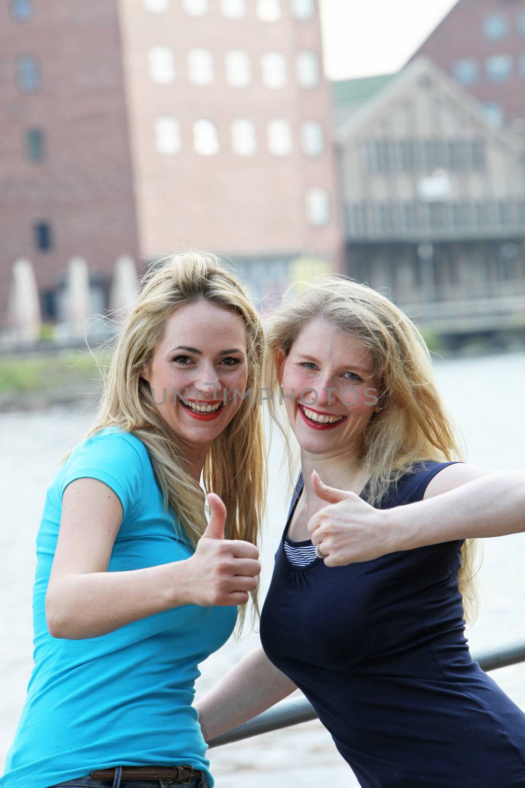 Happy female friends standing alonside an urban waterway giving an enthusiastic thumbs up of approval for their lifestyle Happy female friends standing alonside an urban waterway giving a thumbs up of approval for their lifestyle 