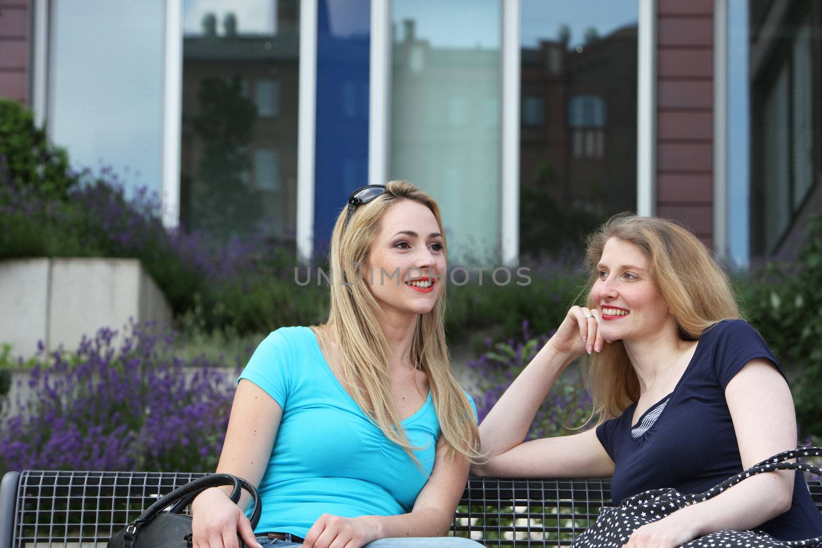 Two attractive young women relaxing in the sunshine on a metal bench chatting in front of a building Two women relaxing in the sunshine on a metal bench chatting in front of a building 