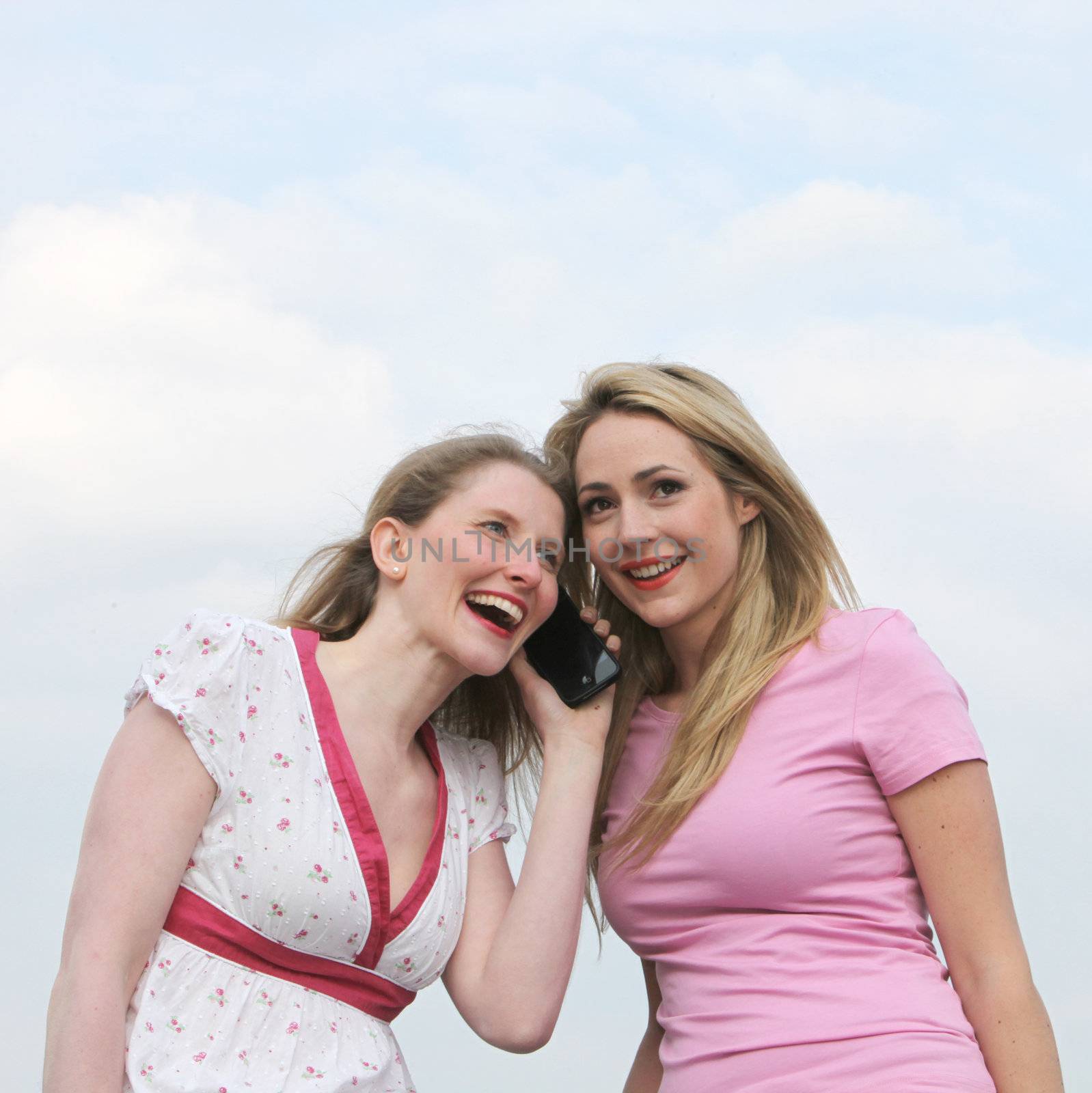 Two attractive woman standing with their heads close together listening to a phone call on the same mobile phone Two attractive woman standing with their heads close together listening to a phone call on the same mobile phone