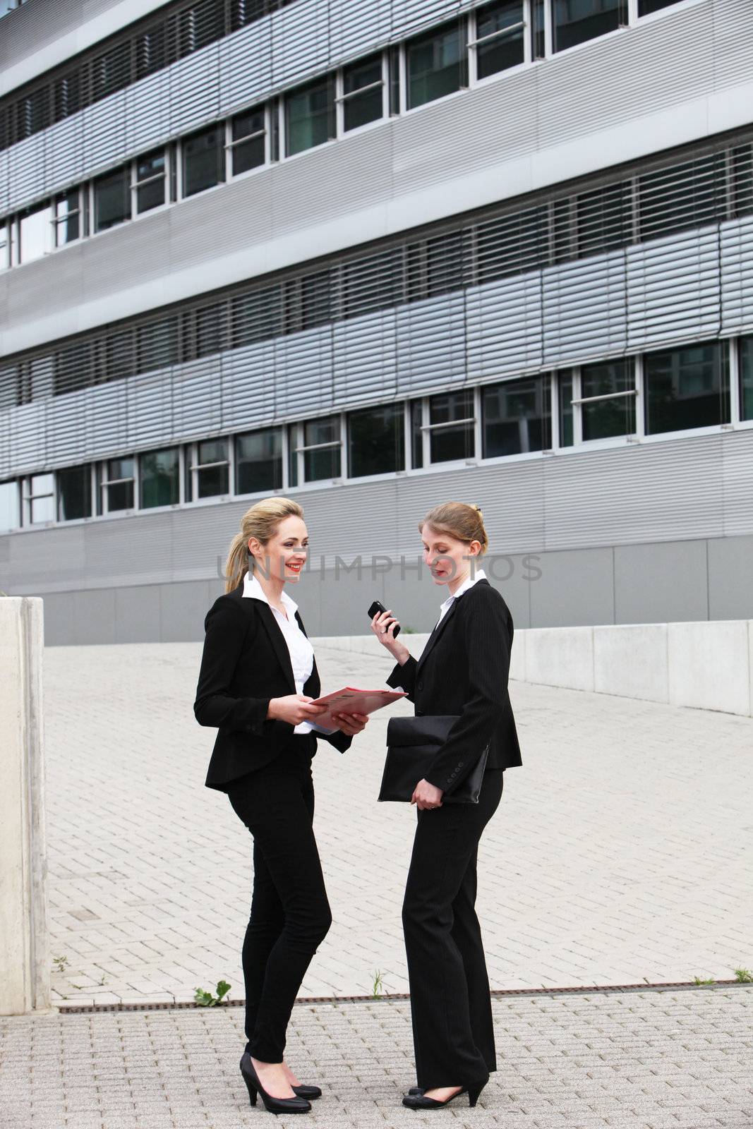 Two stylish female business executives in black slacksuits standing outside an office block in deep conversation 