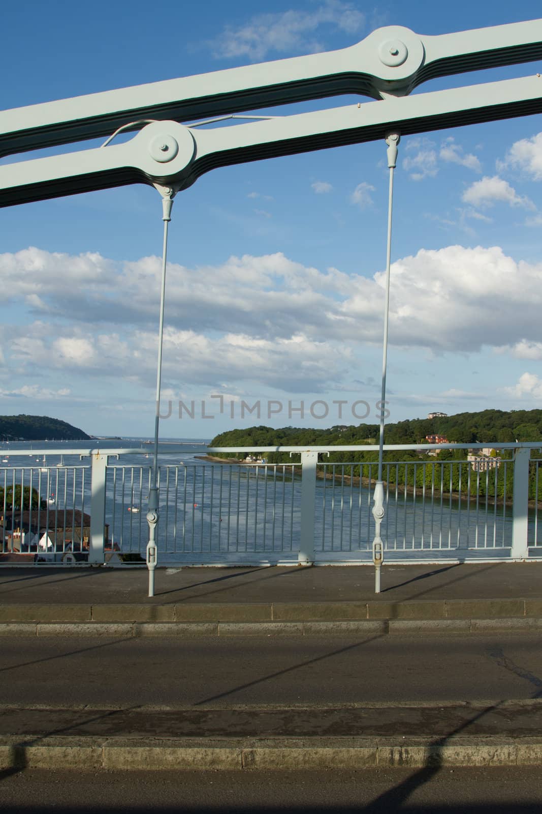 A section of a link chain with cables leading to a road as part of a suspension bridge with water in the background.