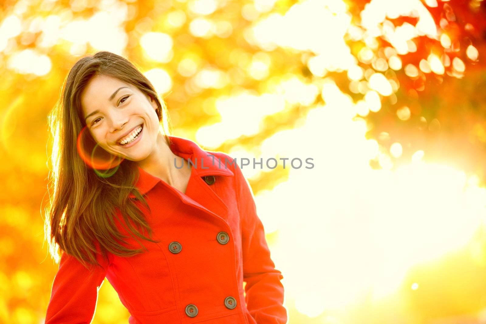 Autumn fashion girl wearing red trench coat in sun flare foliage. Fall woman portrait of happy lovely and beautiful mixed race Asian Caucasian young woman in forest in fall colors.