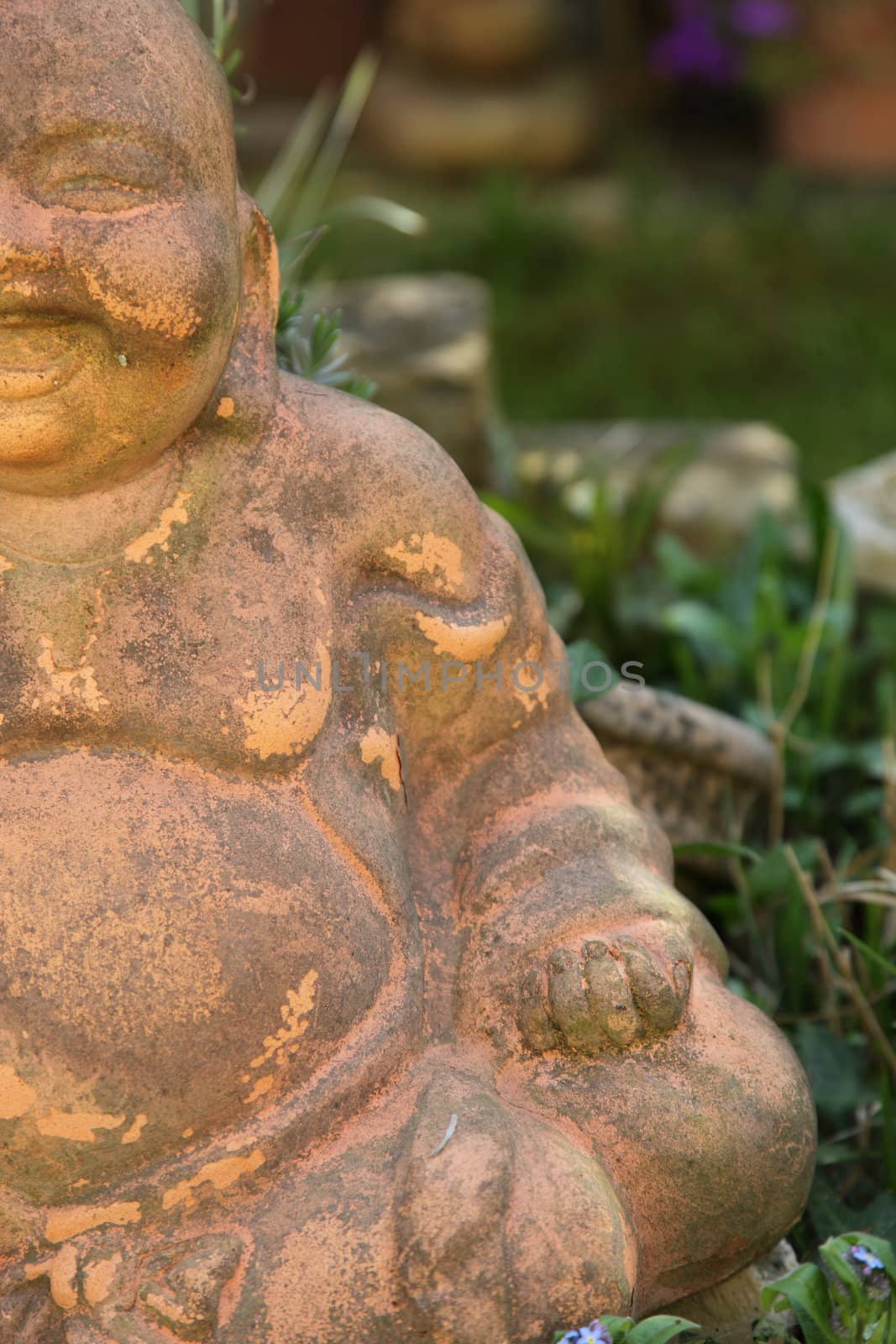 Partial half view of an old terracotta smiling Buddha figurine sitting outdoors amongst greenery