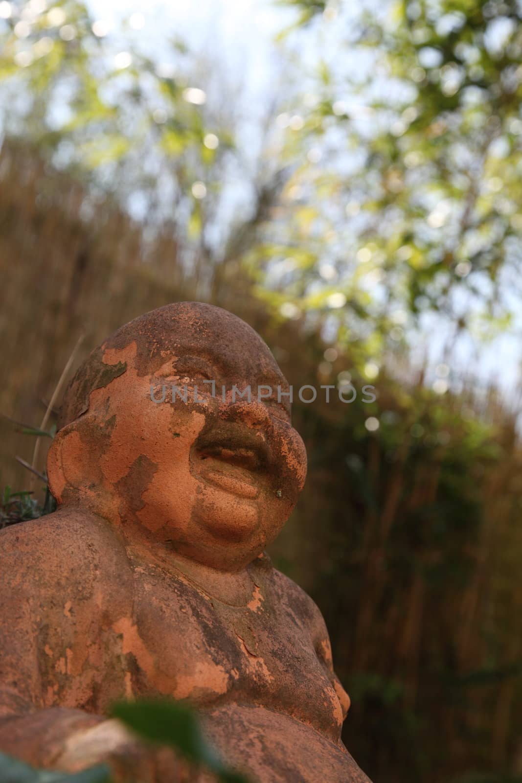 Low angle view of the face of a terracotta smiling Buddha statue outdoors looking up through greenery to blue sky