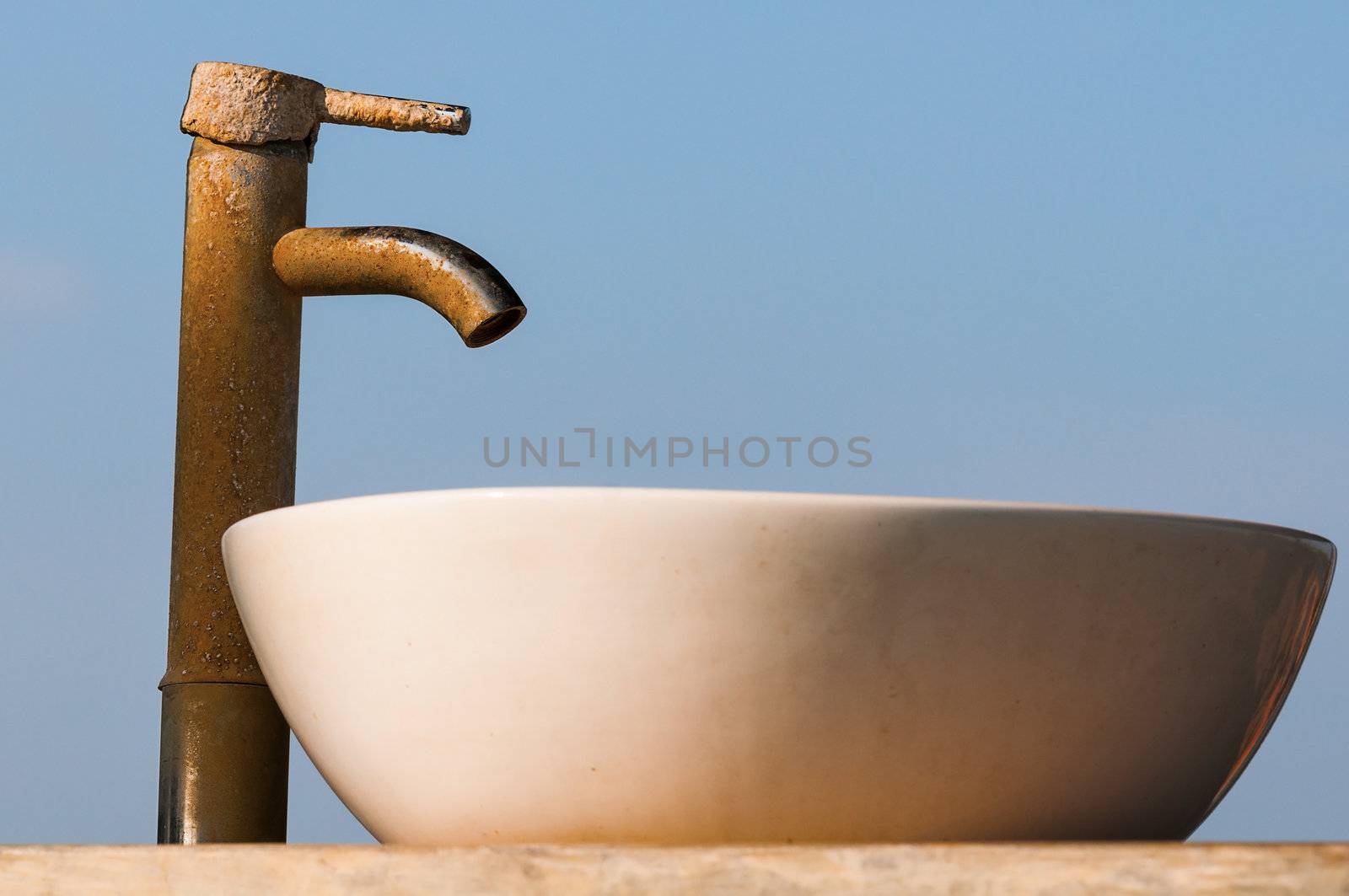 Washbasin and tap covered by limescale with blue sky on background. Focus on the tap. 