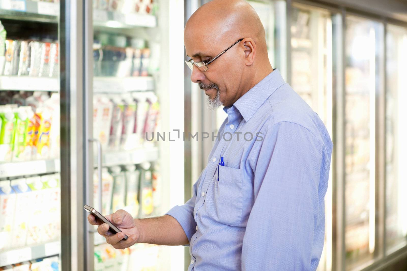 Smiling mature man looking at mobile phone while standing in front of refrigerator in supermarket