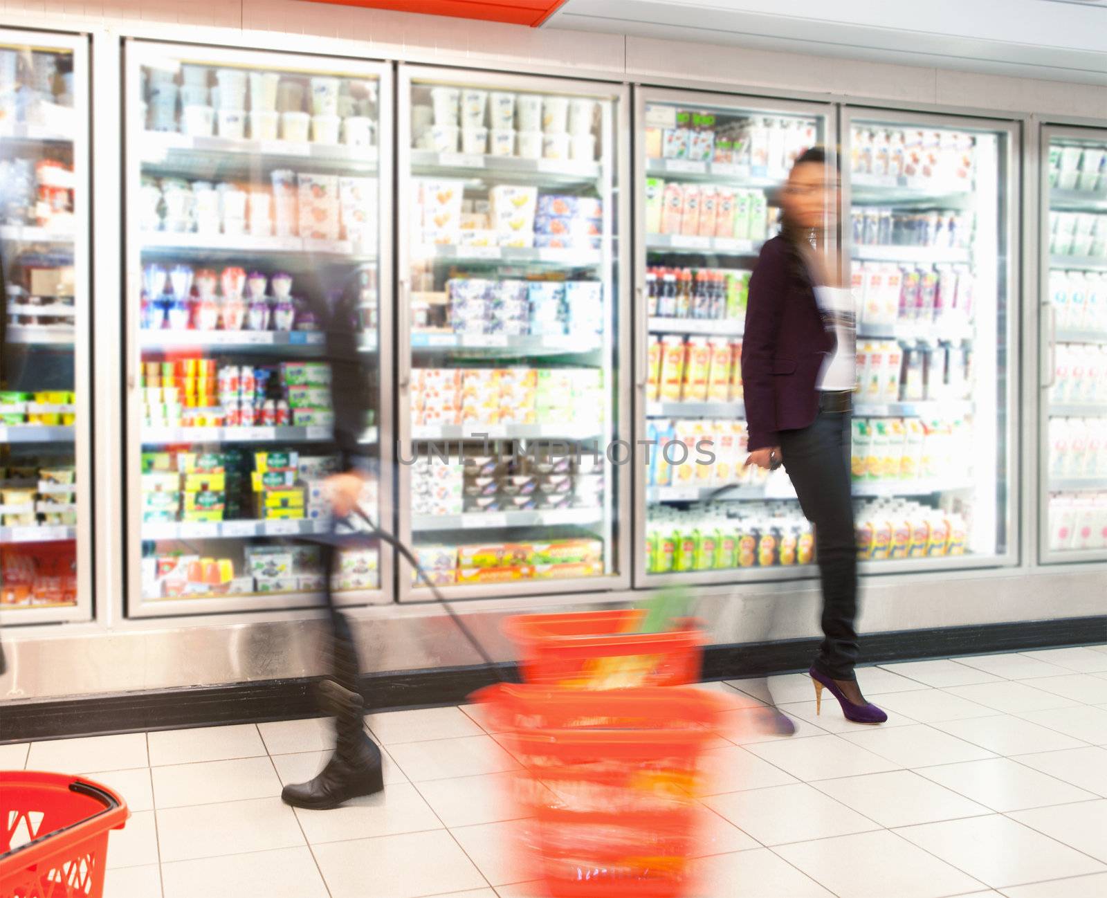 Blurred motion of people walking near refrigerator in shopping centre with baskets