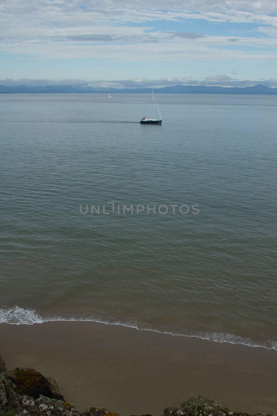 A small wave breaks on a sandy shore and a yacht motors in a green, blue sea with mountains on the horizon.