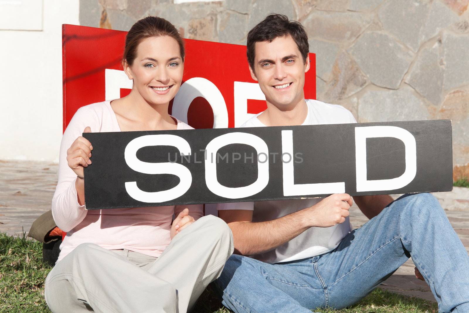 Happy couple sitting against their new house with sold sign