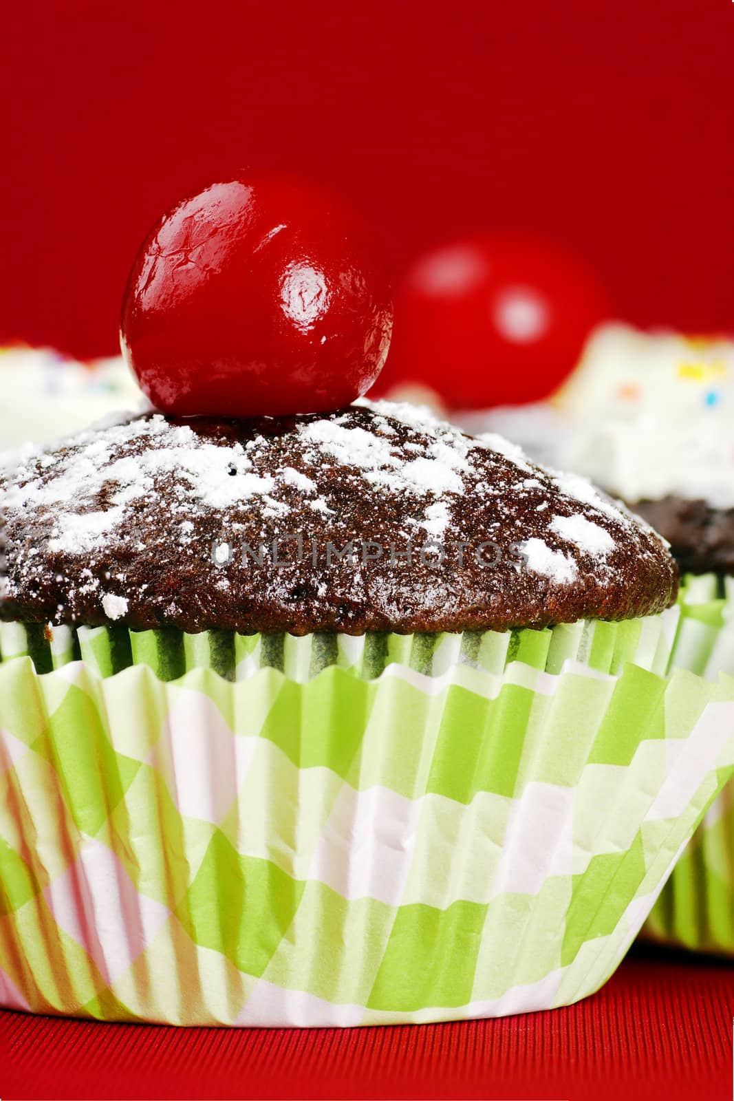 Delicious chocolate cupcakes topped with powdered sugar and maraschino cherry, other desserts with frosting and candies at the back, over red, stacked focus.