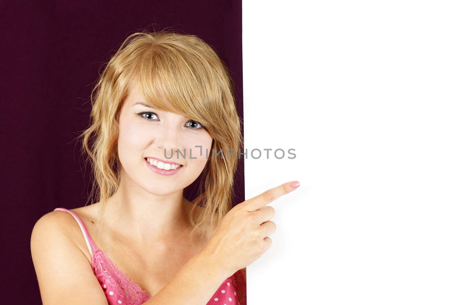 Cute and friendly smiling young blond teenager girl holding and pointing at a white blank sign, placard or billboard.