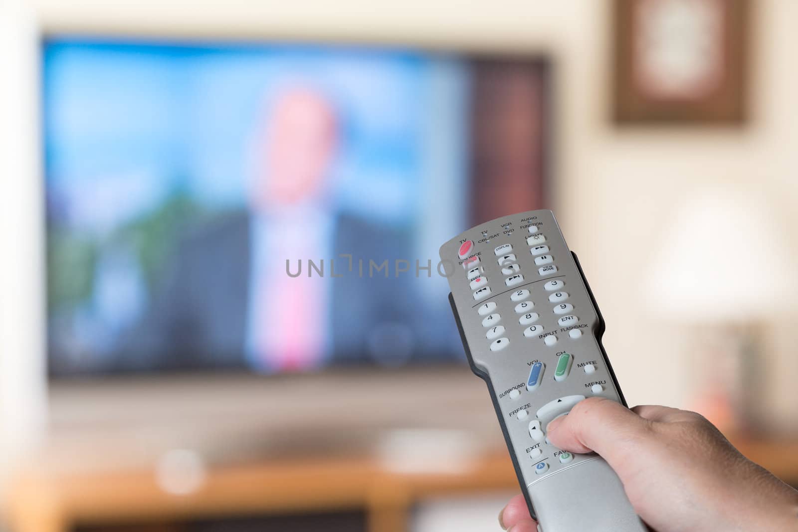 Silver modern TV remote control being pressed by thumb with out of focus screen background