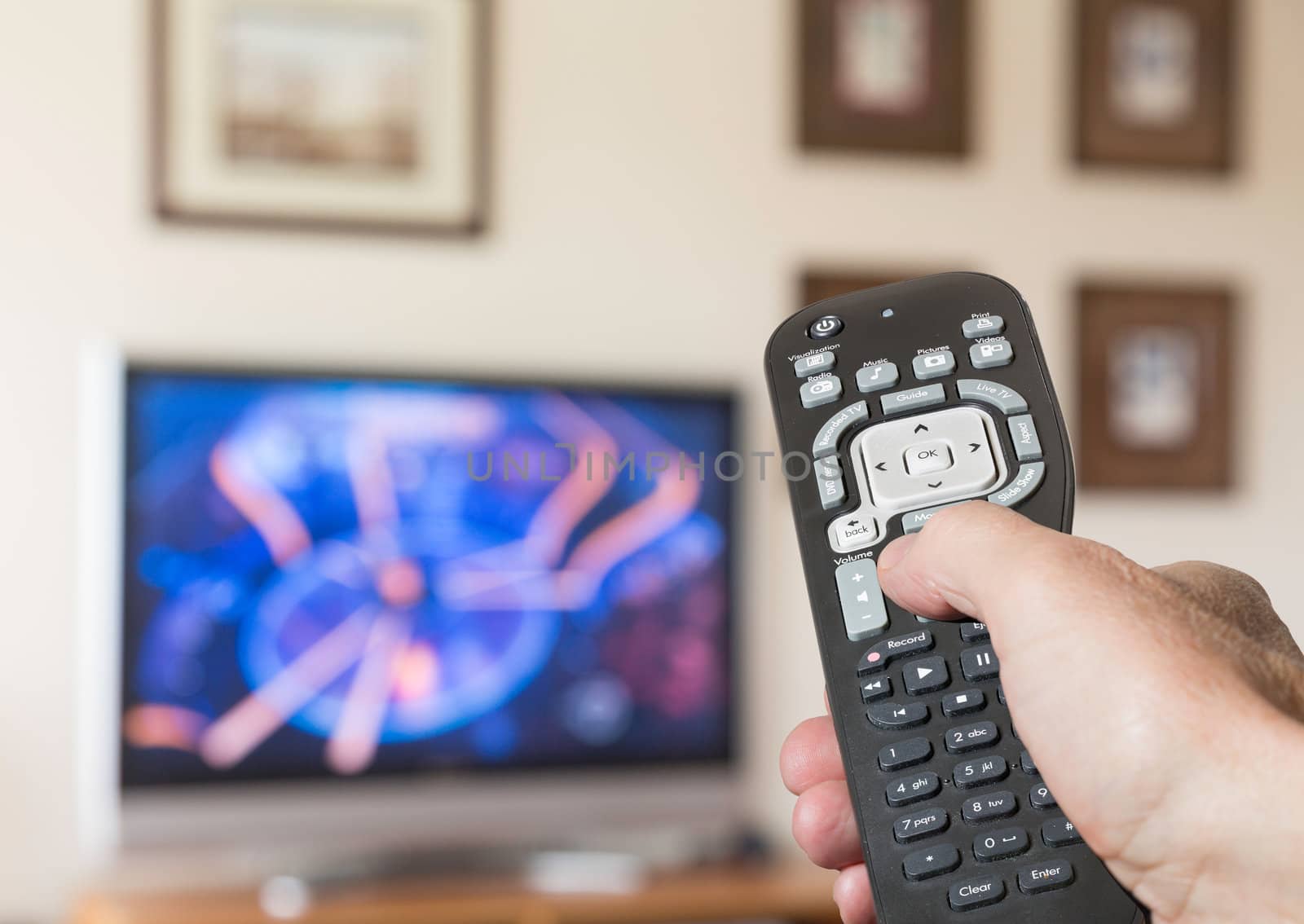 Black modern TV remote control being pressed by thumb with out of focus screen background