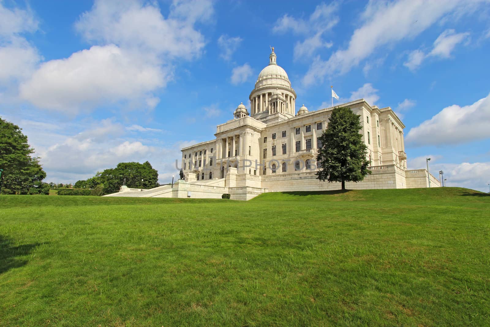 Wide-angle view of the front of the Rhode Island state capitol building as it sits on Capitol Hill in Providence with bright blue sky and white clouds in the background. The building is covered with white Georgia marble and was constructed from 1895 to 1904.