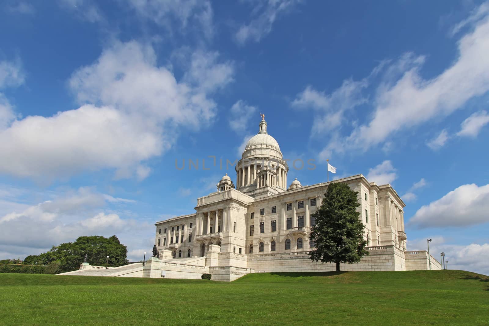 Wide-angle view of the front of the Rhode Island state capitol building as it sits on Capitol Hill in Providence with bright blue sky and white clouds in the background. The building is covered with white Georgia marble and was constructed from 1895 to 1904.