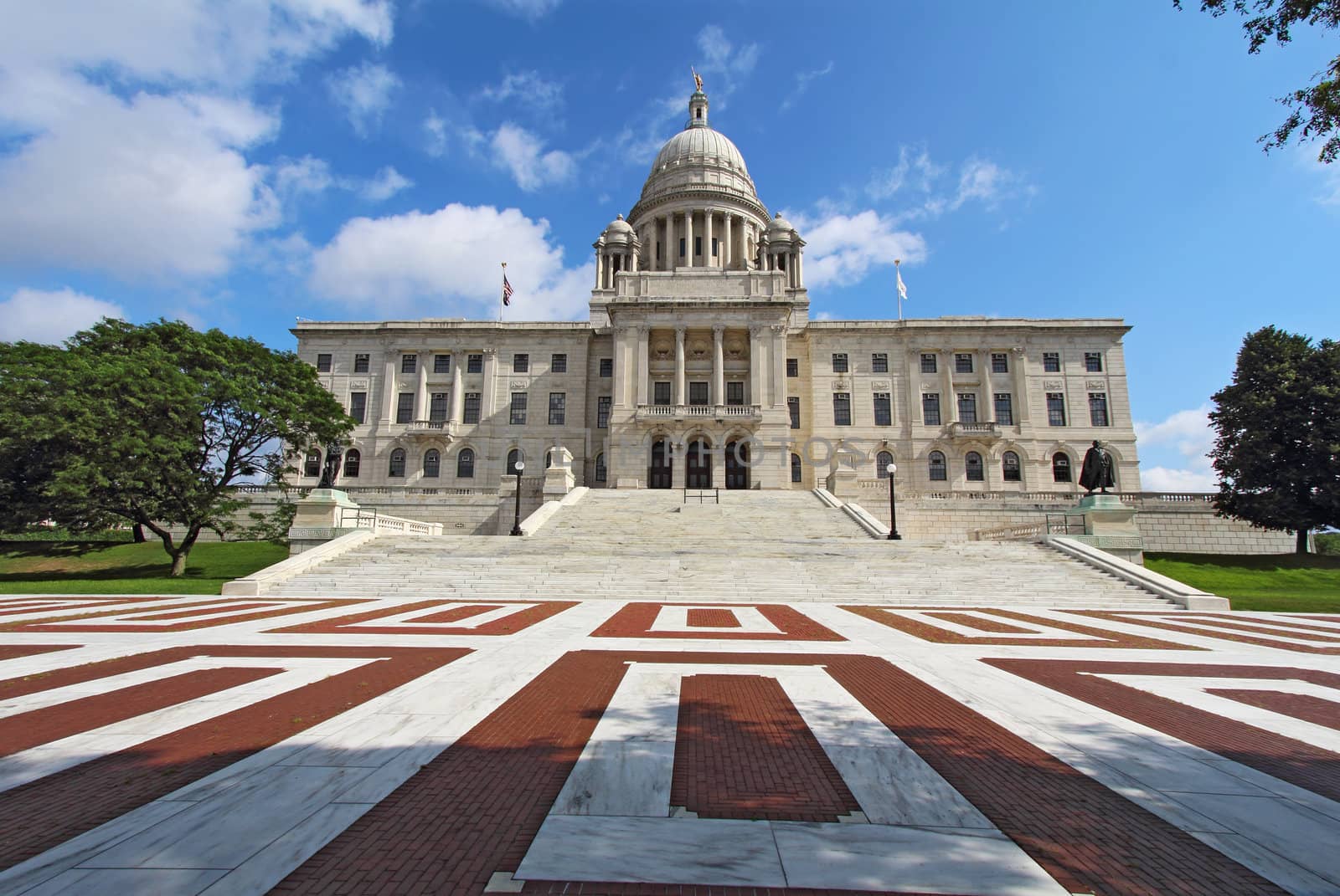 Wide-angle view of the front of the Rhode Island state capitol building with red and white brickwork in Providence with bright blue sky and white clouds in the background. The building is covered with white Georgia marble and was constructed from 1895 to 1904.