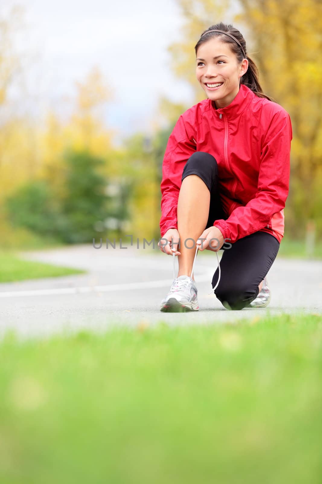 Runner woman tying running shoes outside in fall. Beautiful young fitness model smiling happy in casual jogging clothing.