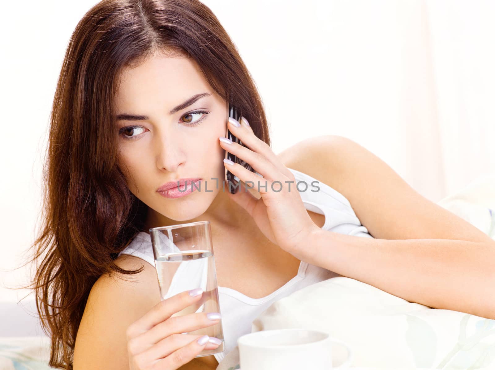 Pretty woman making phone call and holding glass of water in bed