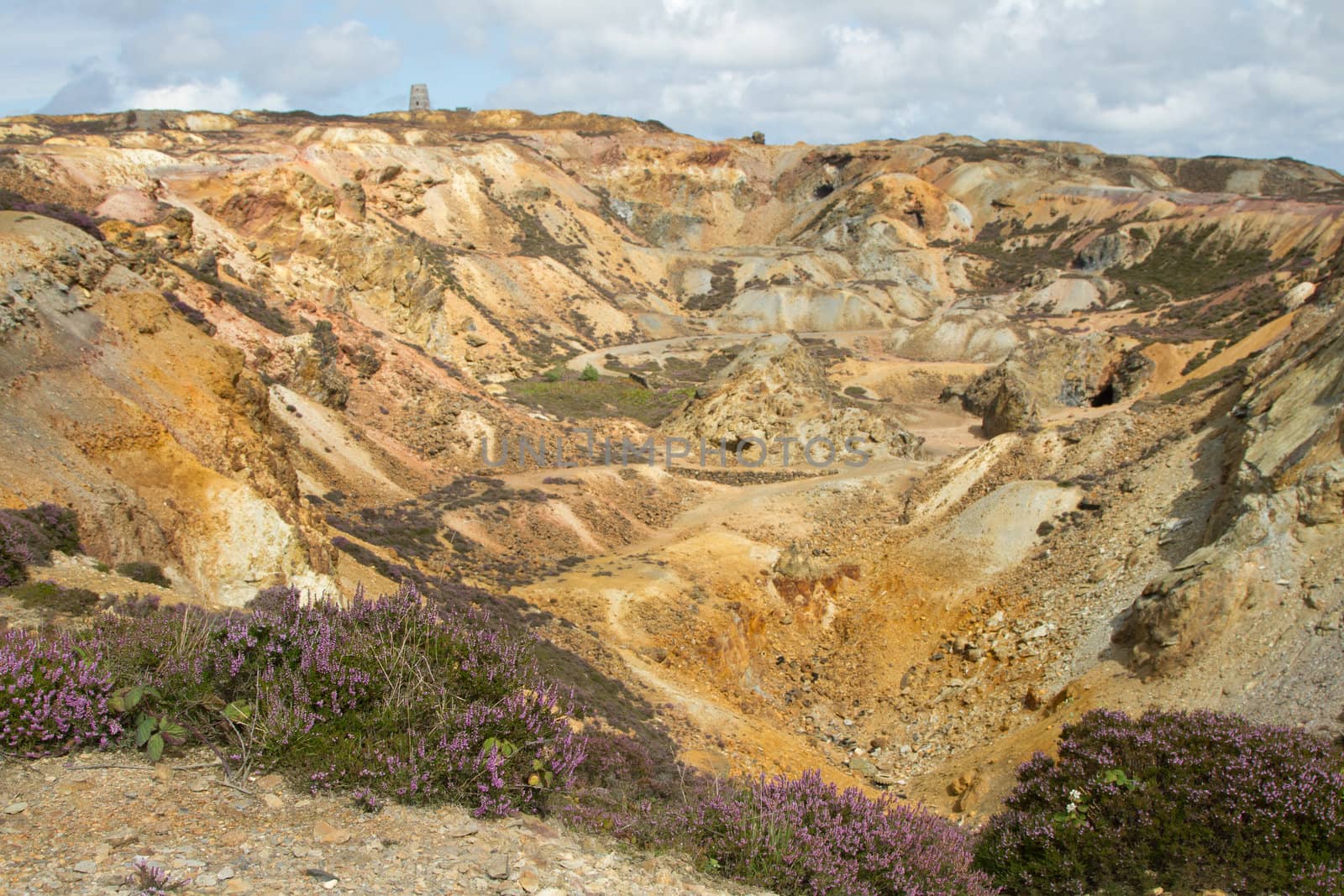 An old opencast copper mine excavation with multicoloured metal ore rock and a building in the distance against a cloudy sky.