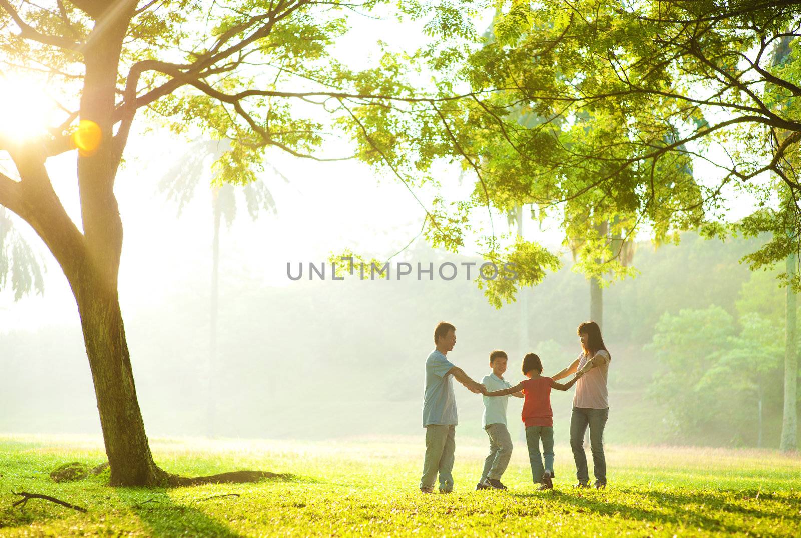 Happy Asian family holding hands in a circle and running over green lawn