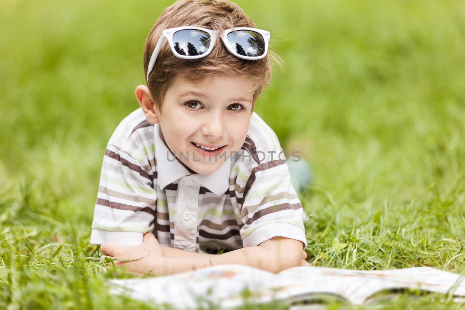 Beauty smiling child boy in sunglasses reading book outdoor on green grass field