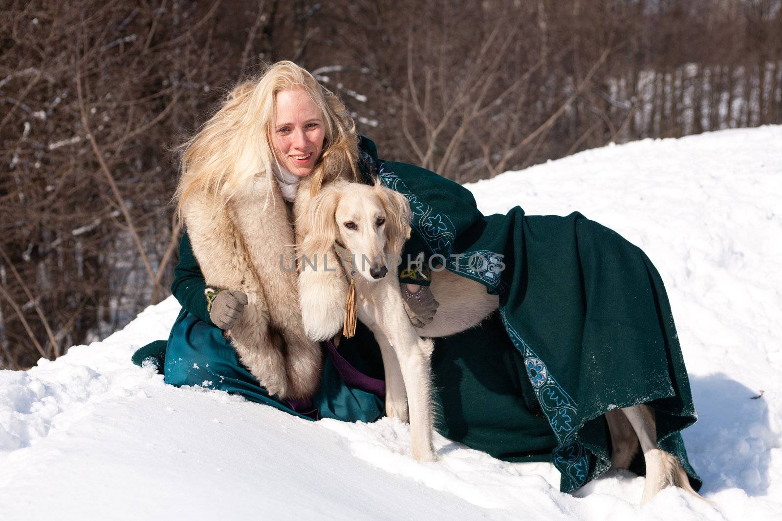 A blonde girl and a standing white saluki on snow
