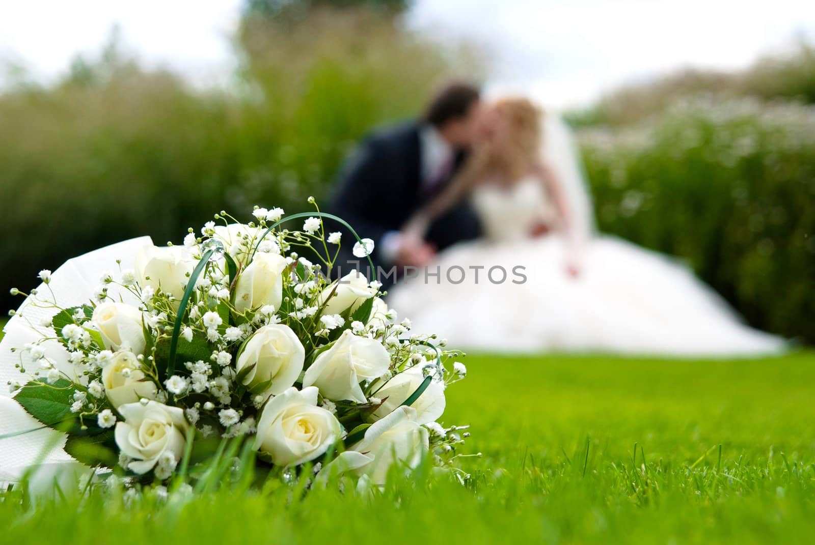 Bridal bouquet of white roses on a green meadow and blurred newlyweds