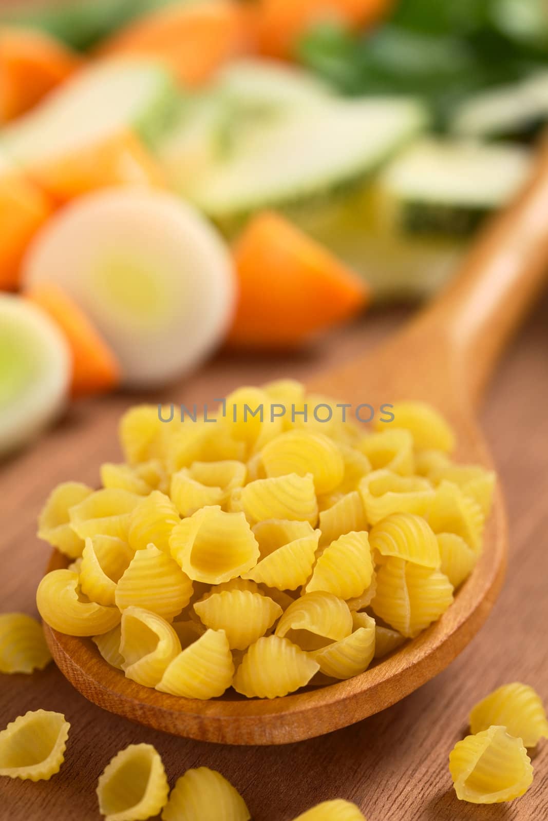 Raw shell pasta or conchiglioni on wooden spoon with raw vegetables (carrot, leek, zucchini) in the back (Selective Focus, Focus one third into the pasta on the spoon) 