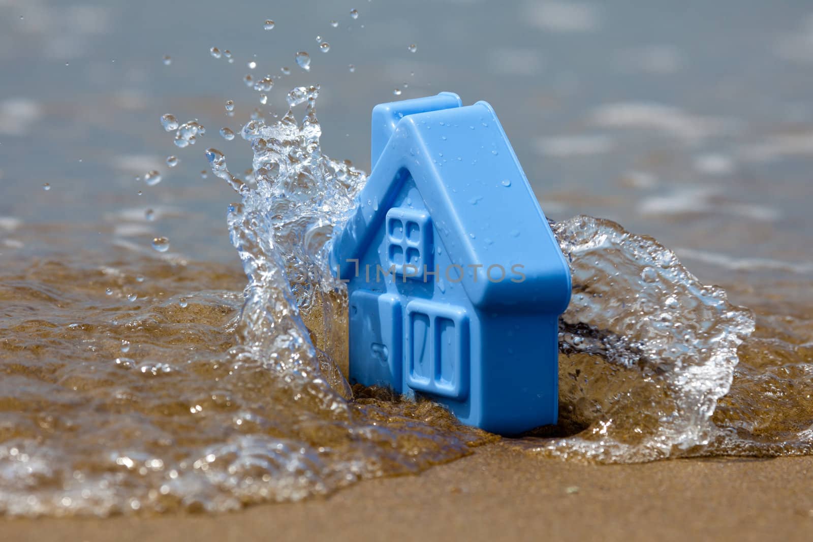 Blue plastic toy house on the sand covered with the waves, forming the spray - a metaphor for the sudden flooding