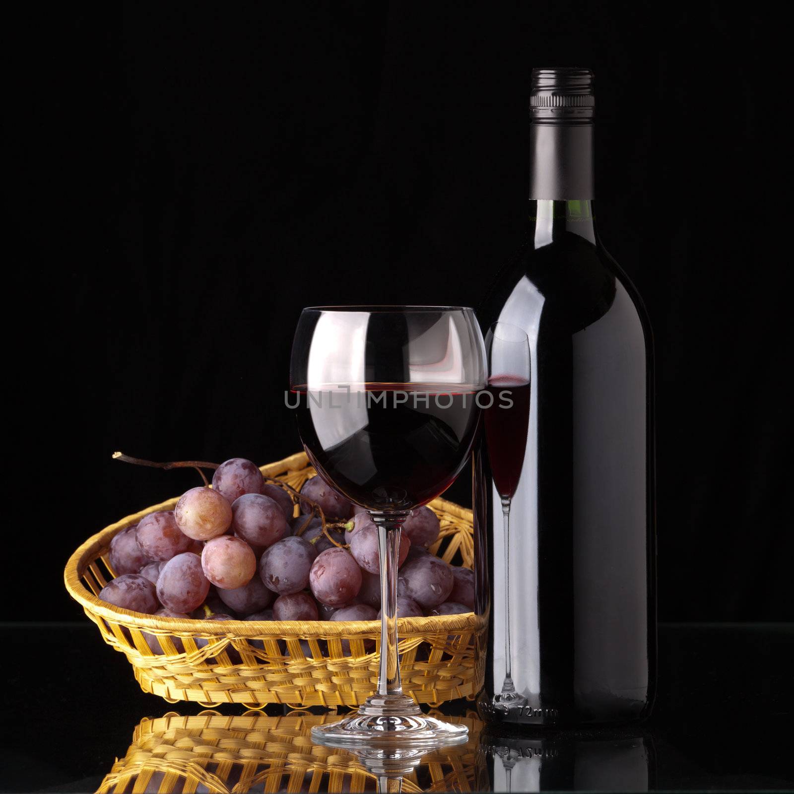 A full bottle of red wine, a glass of wine and black grapes in a wicker basket on a black background