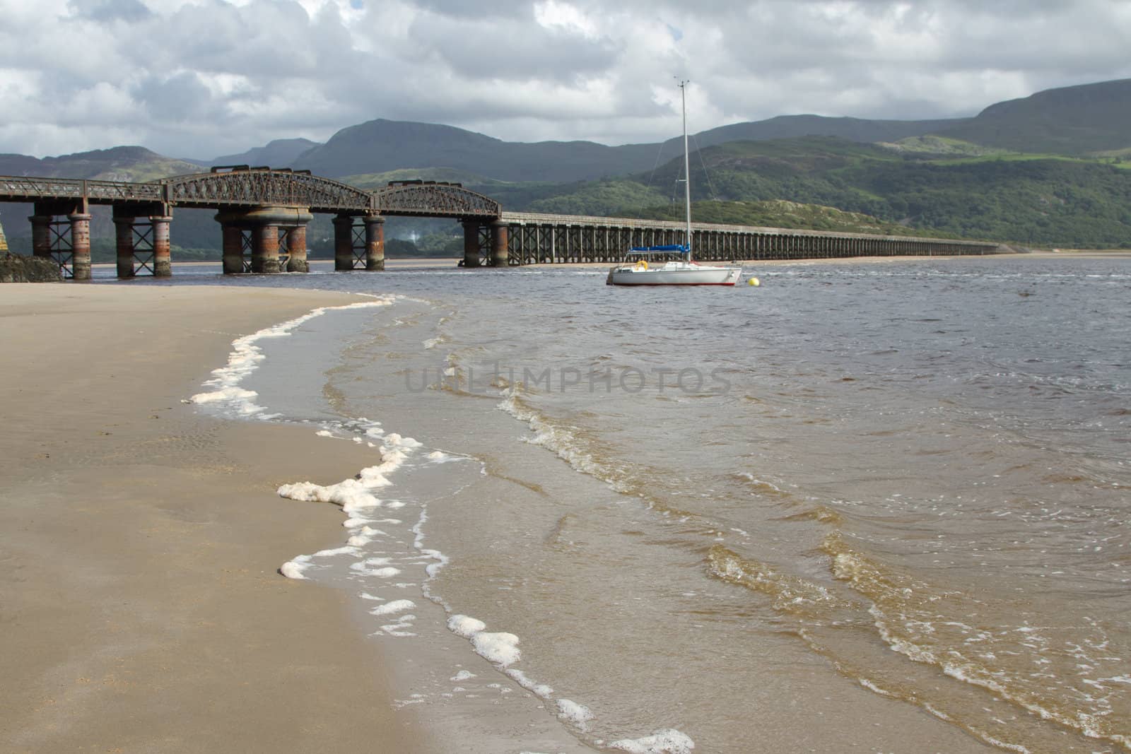 A railway bridge spans an estuary with the sea up on the sand and a is moored in the water backed by dark mountains.