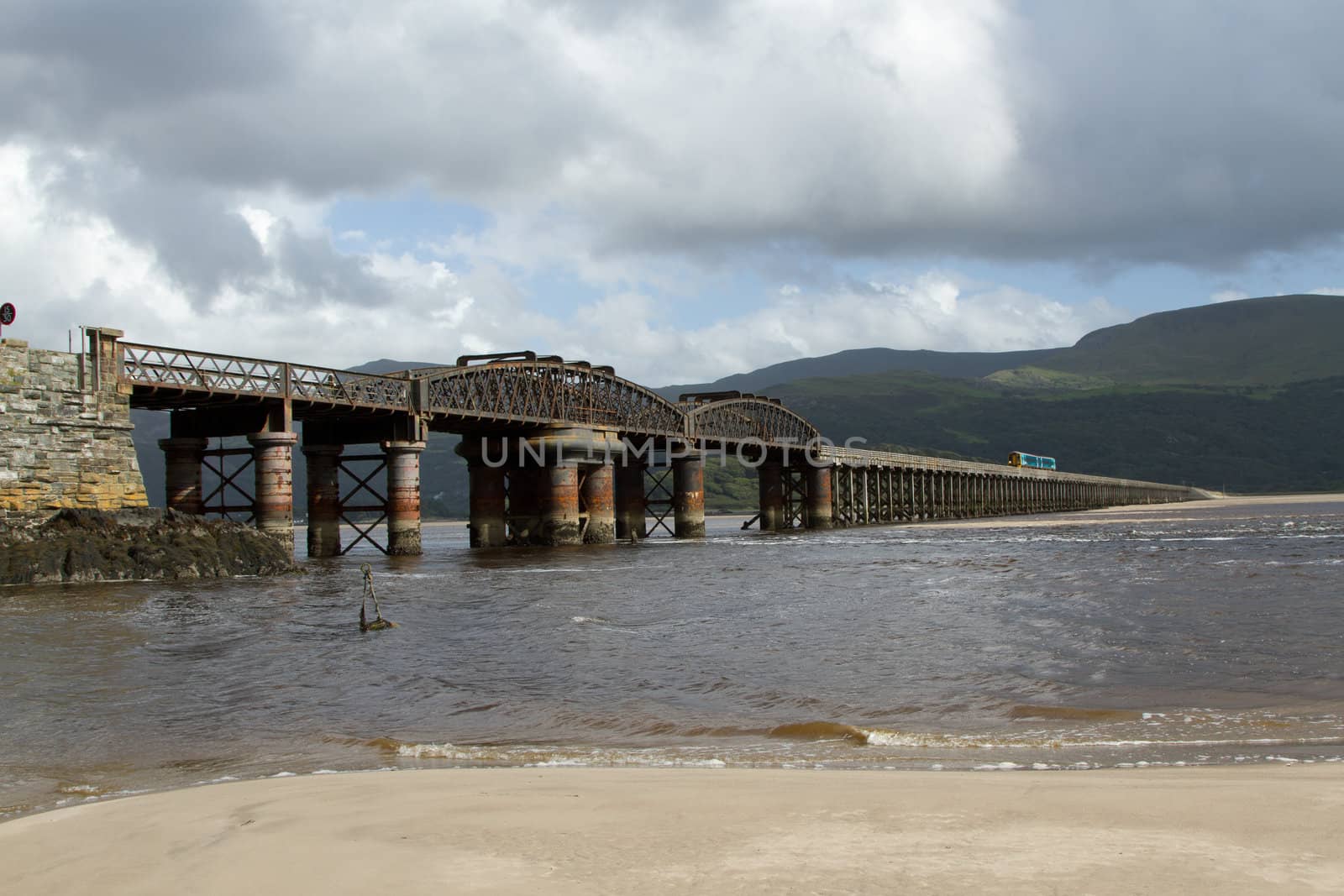 A railway bridge spans an estuary with the sea up on the sand and a train in the distance backed by dark mountains.