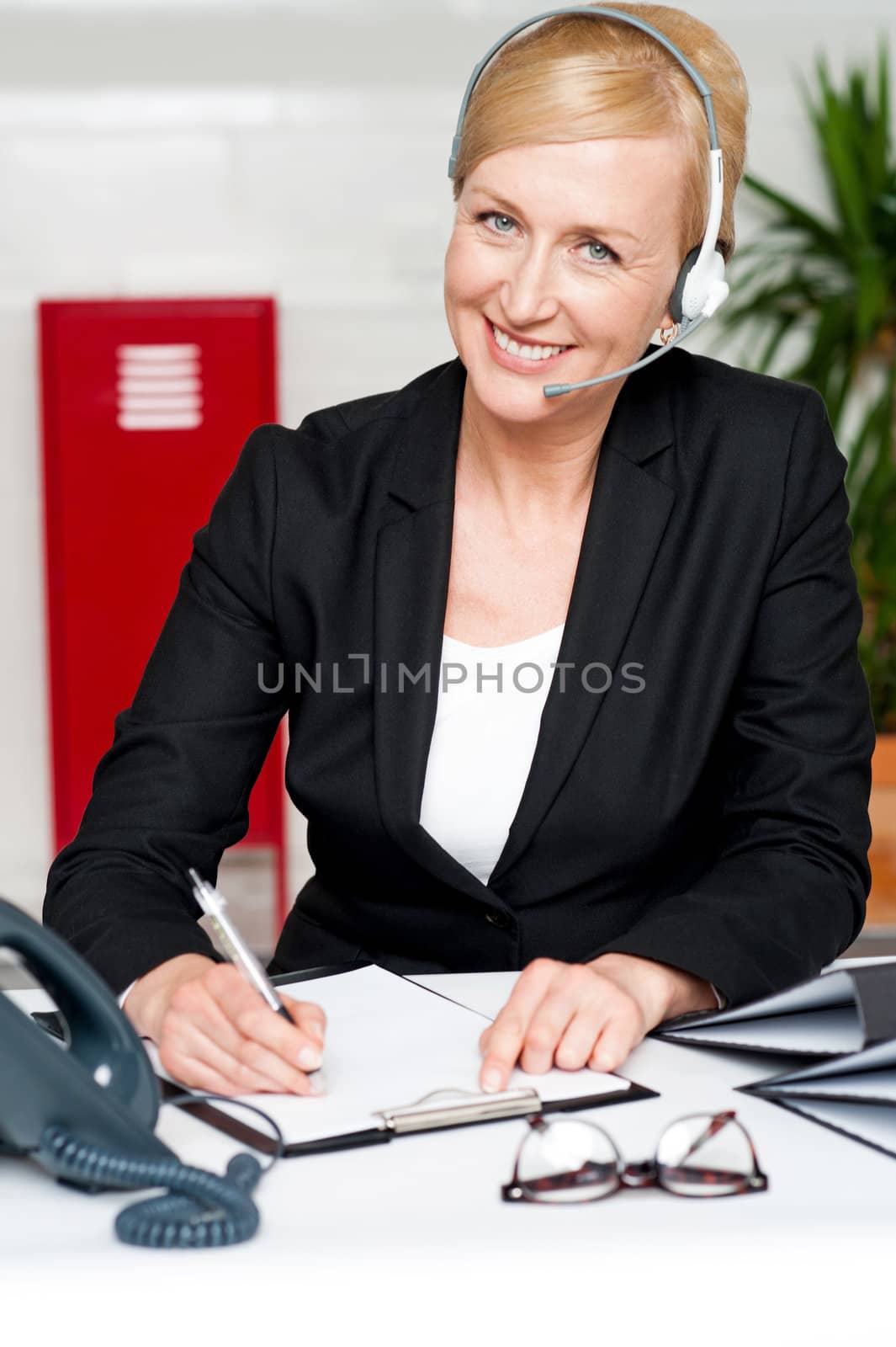 Smiling woman wearing headset writing on notepad sitting in office