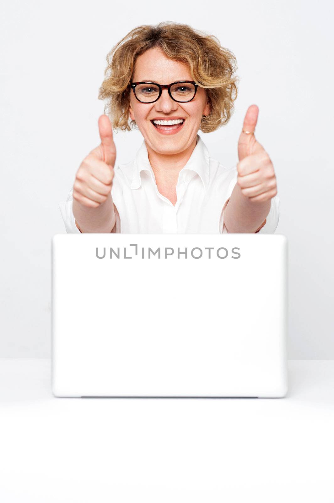 Joyful senior executive showing double thumbs up to camera with laptop kept on desk