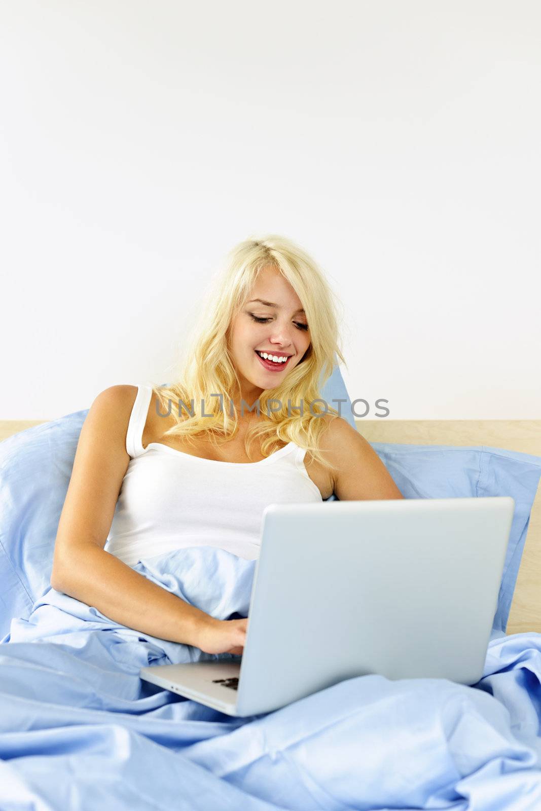 Smiling blonde woman using laptop computer at home sitting in bed