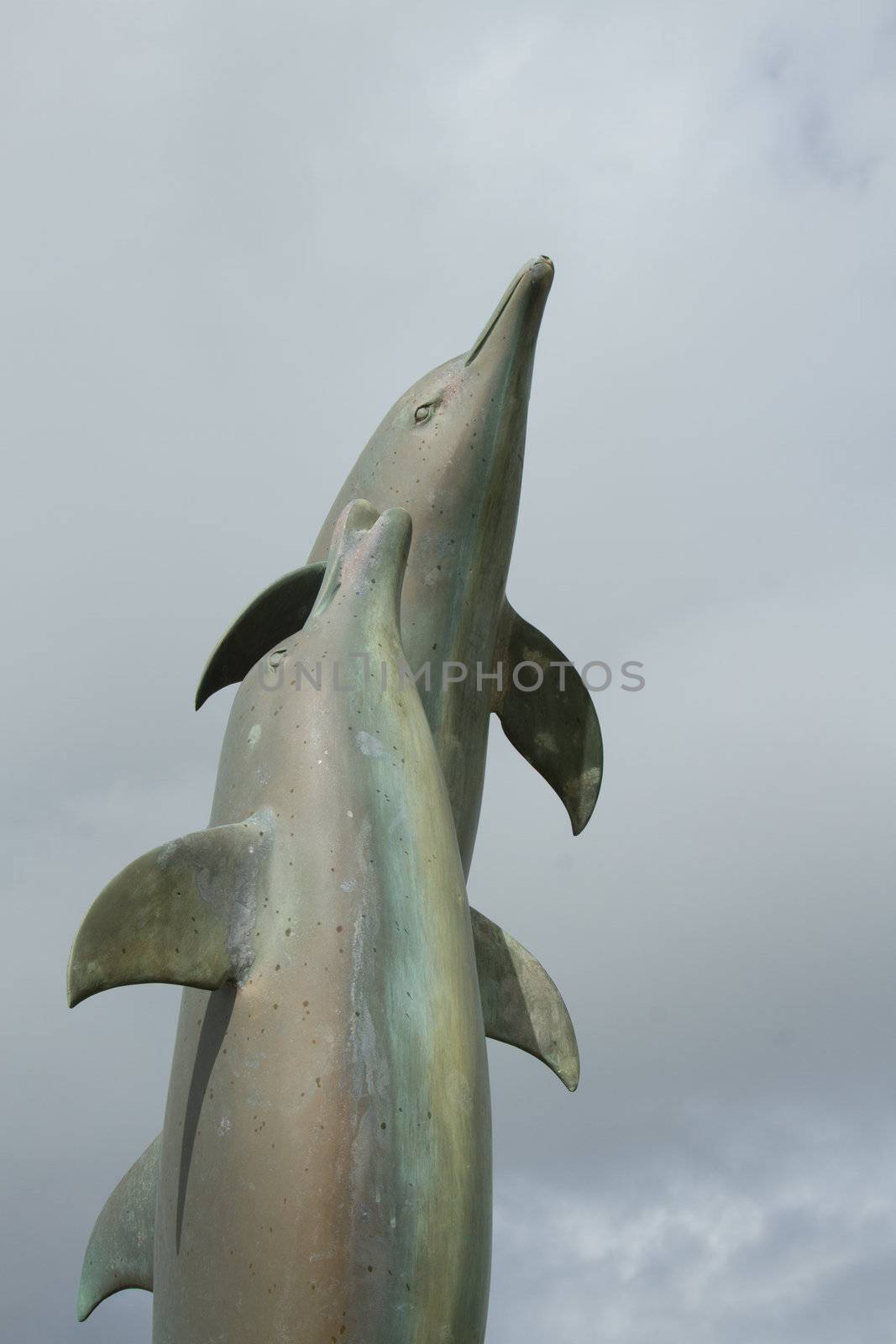 A statue showing a pair of metal moulded dolphins jumping up towards a cloudy sky.