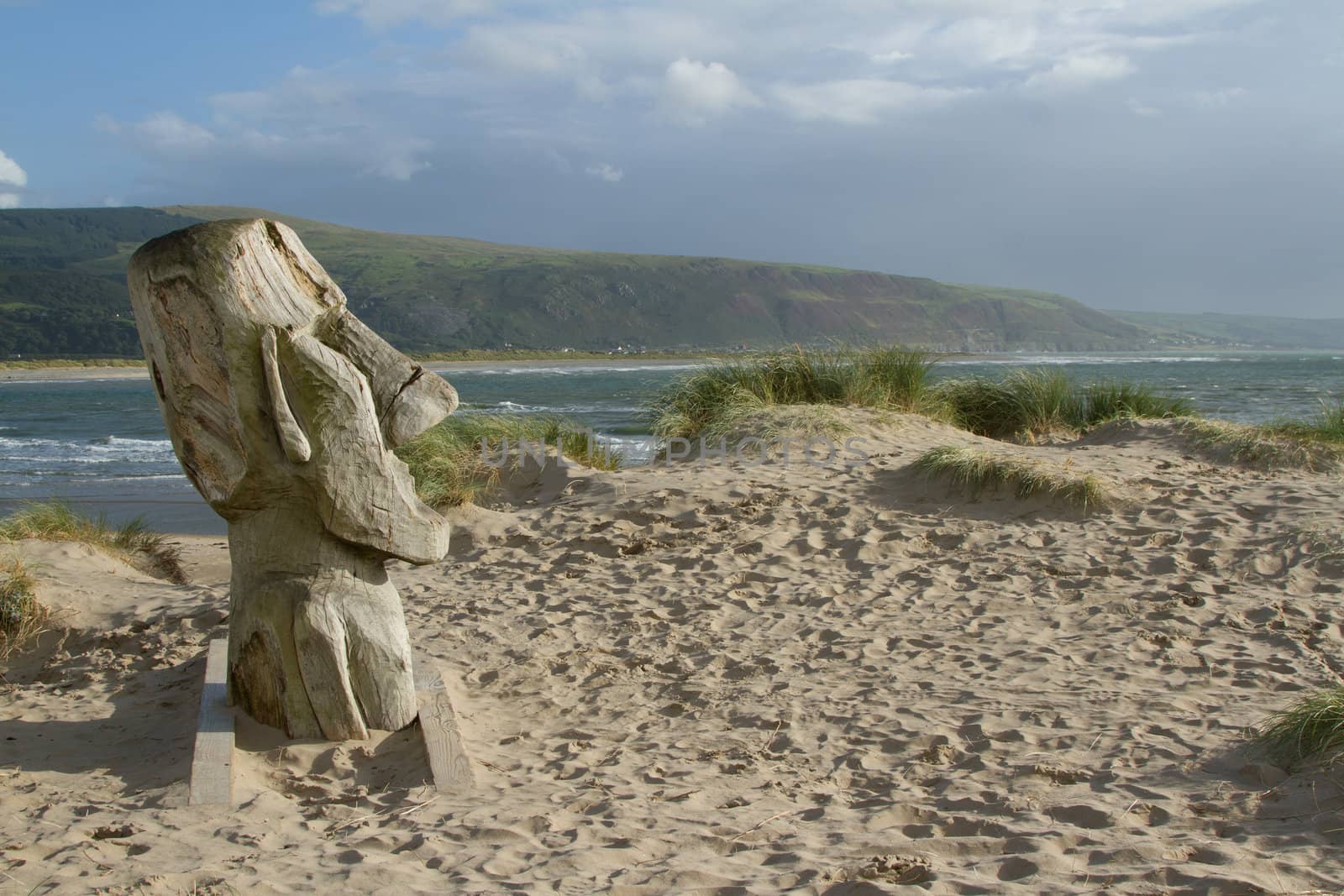 The face of a man carved into a section of wood placed onto a sand dune with grasses, looking out over the sea and distant hills.