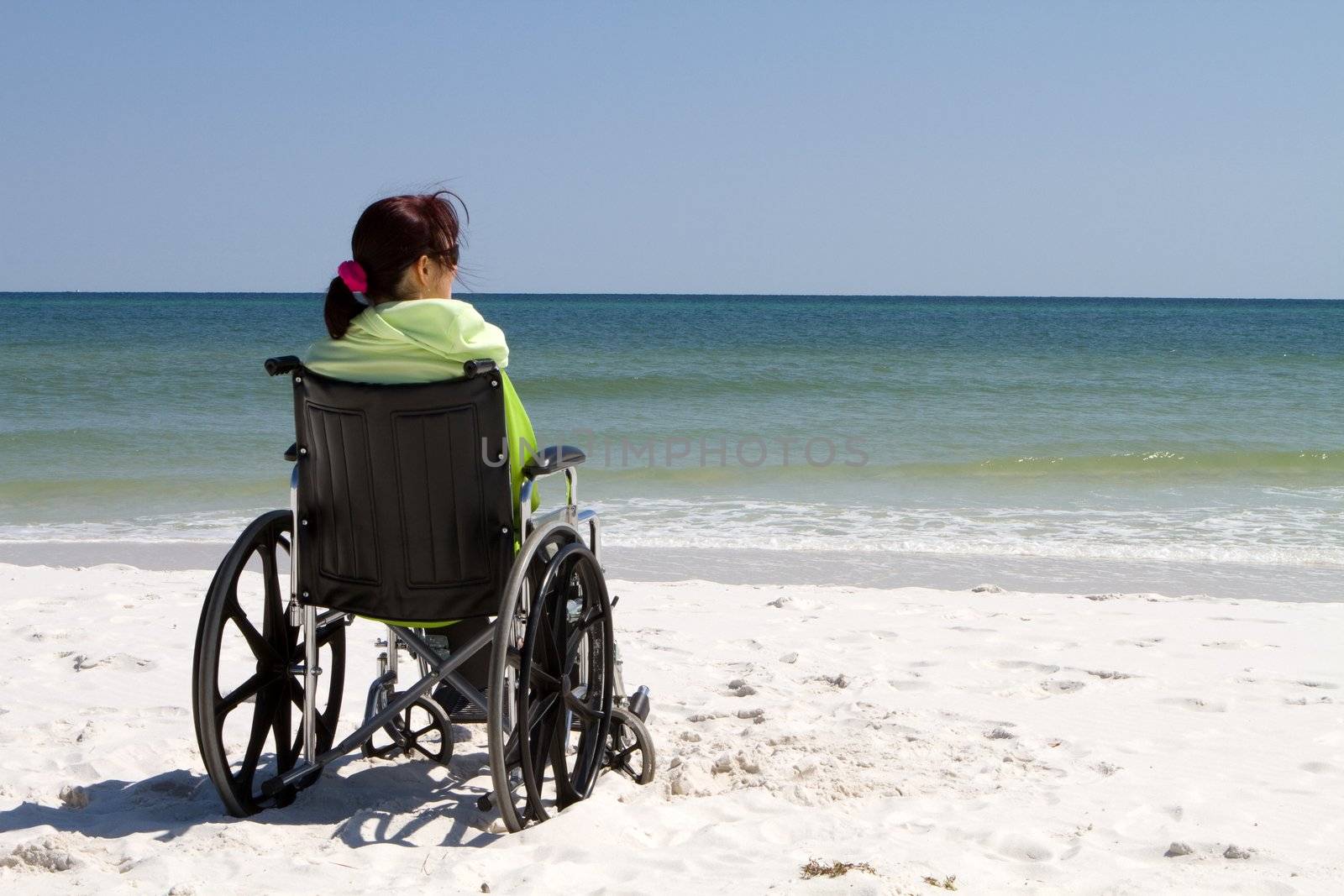 Disabled woman sits alone in a wheelchair on a sandy beach watching the ocean waves.