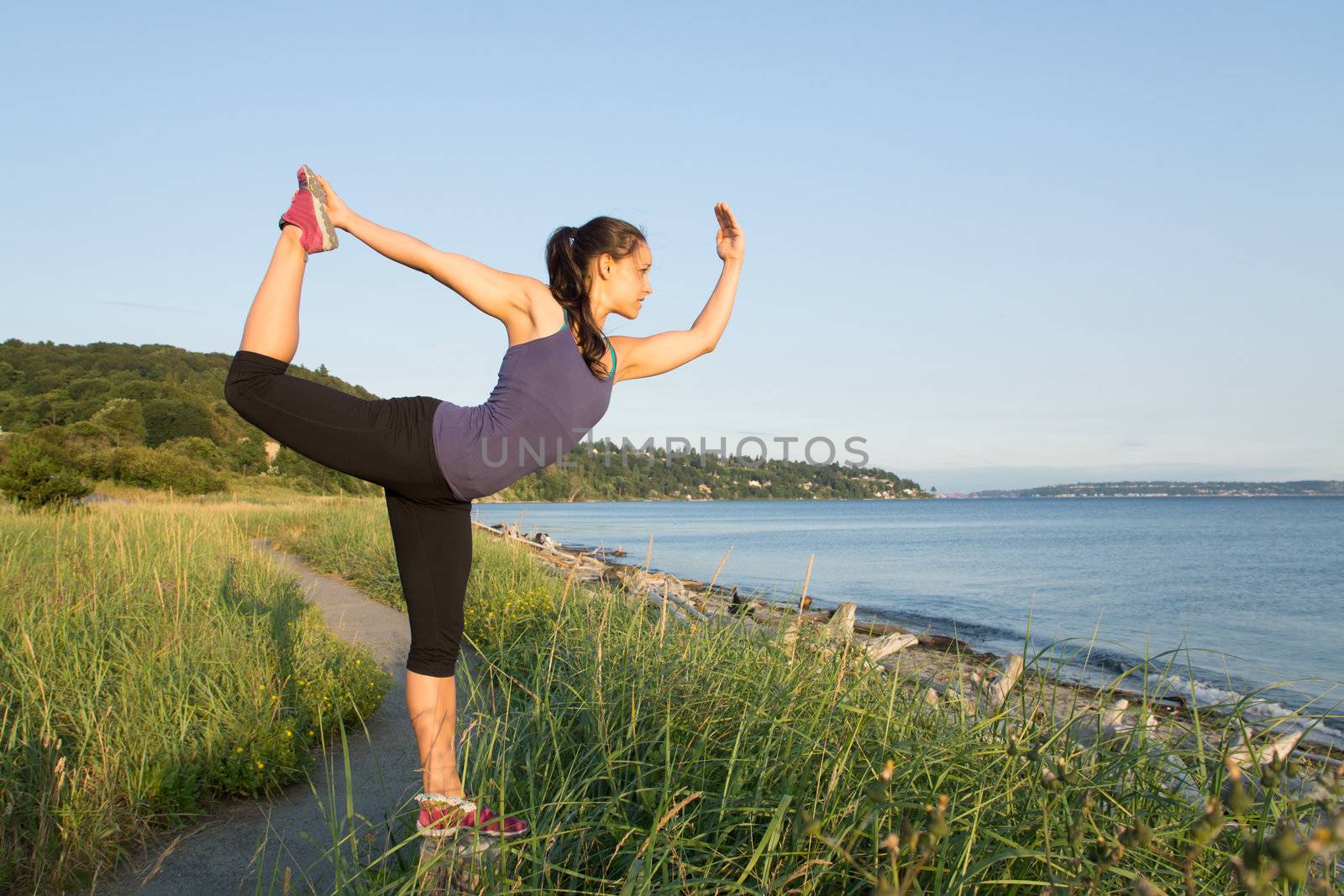 Young women balancing on trail post in Natarajasana pose with shoreline and clear sky in baclgroud.