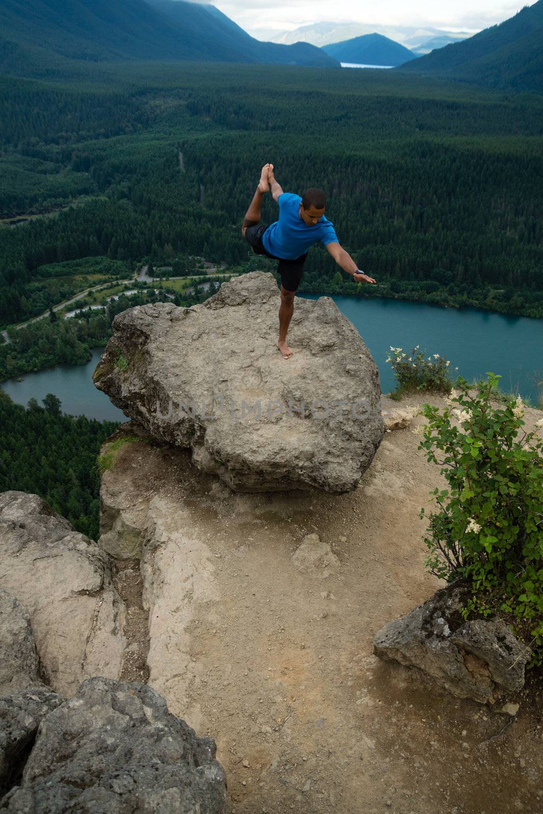 Young man in yoga pose on top of mountain with beautiful vista in background.