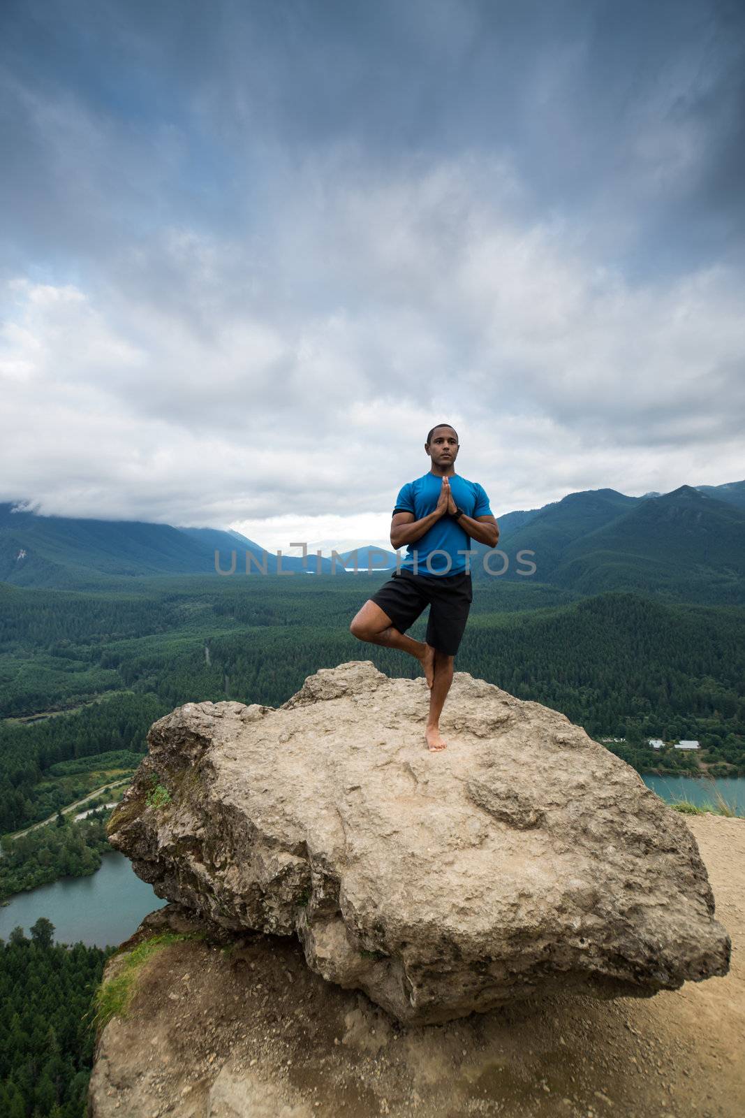 Young man in yoga pose on top of mountain with beautiful vista in background.