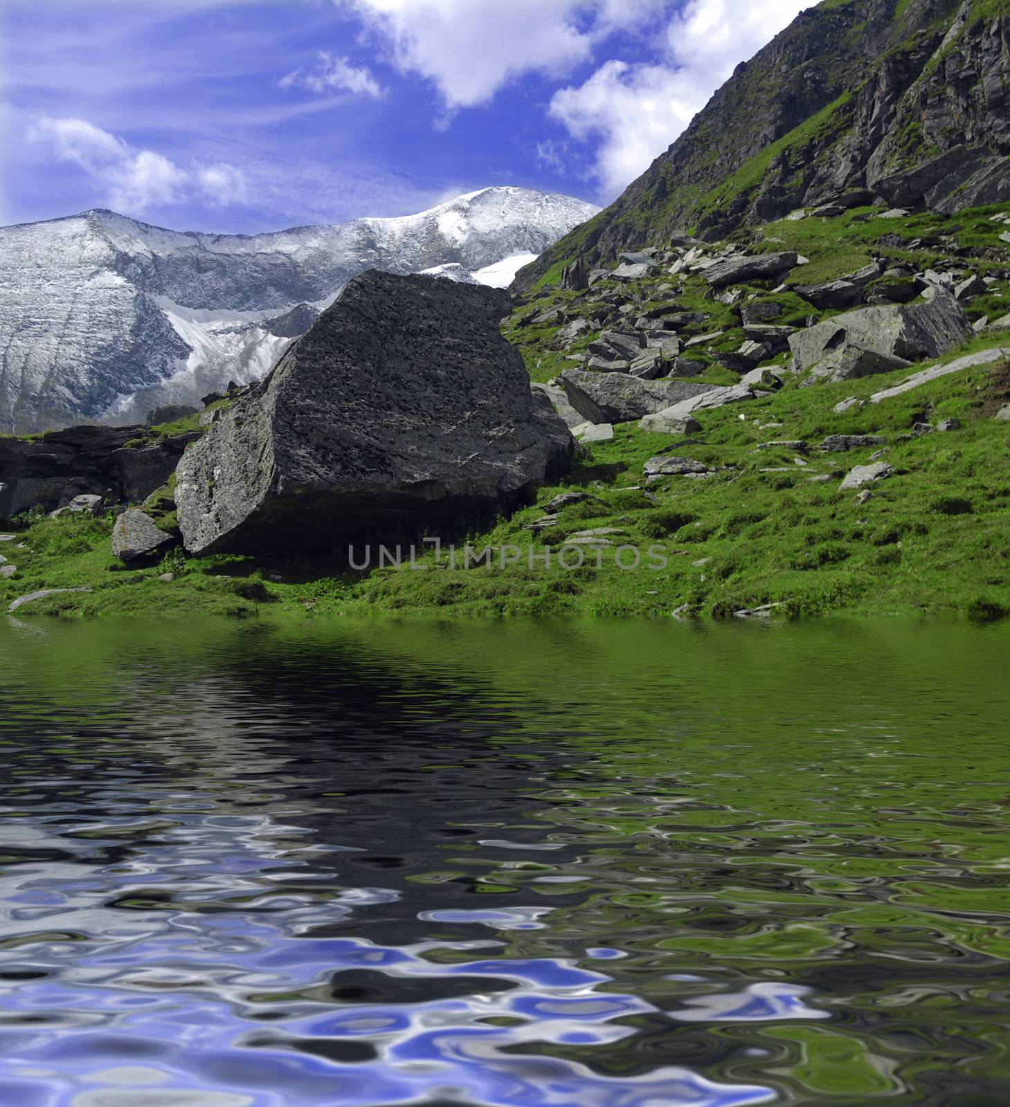 Beautiful Alpine landscape with mountains covered in snow and big boulder  on the grass reflectiong in the lake