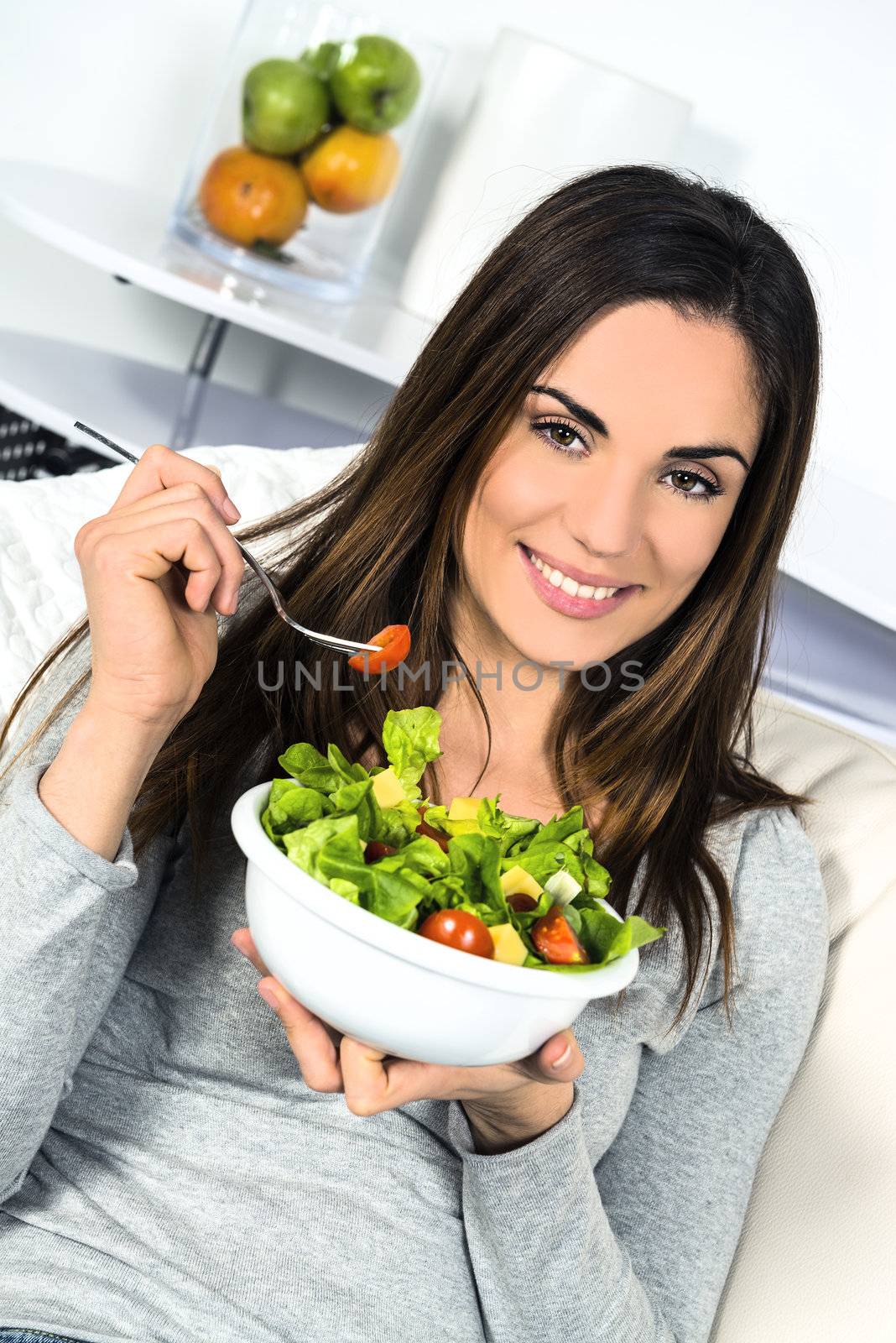 Woman eating salad. Beautiful healthy smiling Caucasian woman enjoying a fresh healthy salad sitting in sofa looking up. High angle view with copy space on modern interior. 