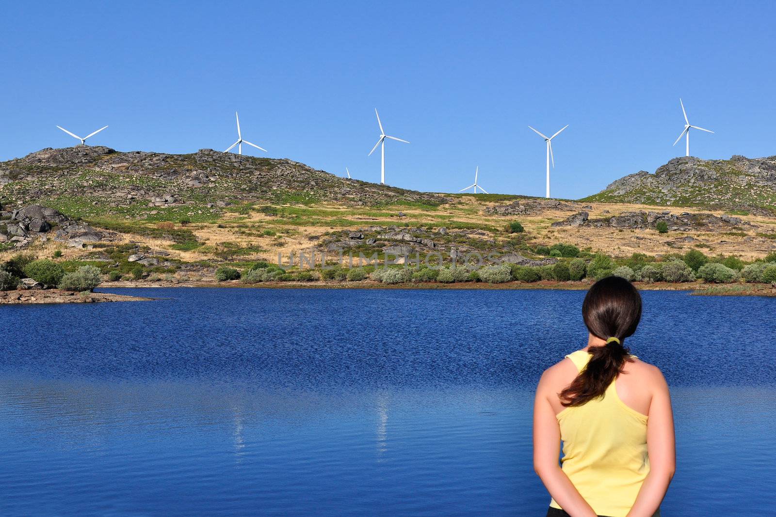 Woman looking at a wind mill across a lake. Focus is on the wind turbines and the woman is slightly out of focus.