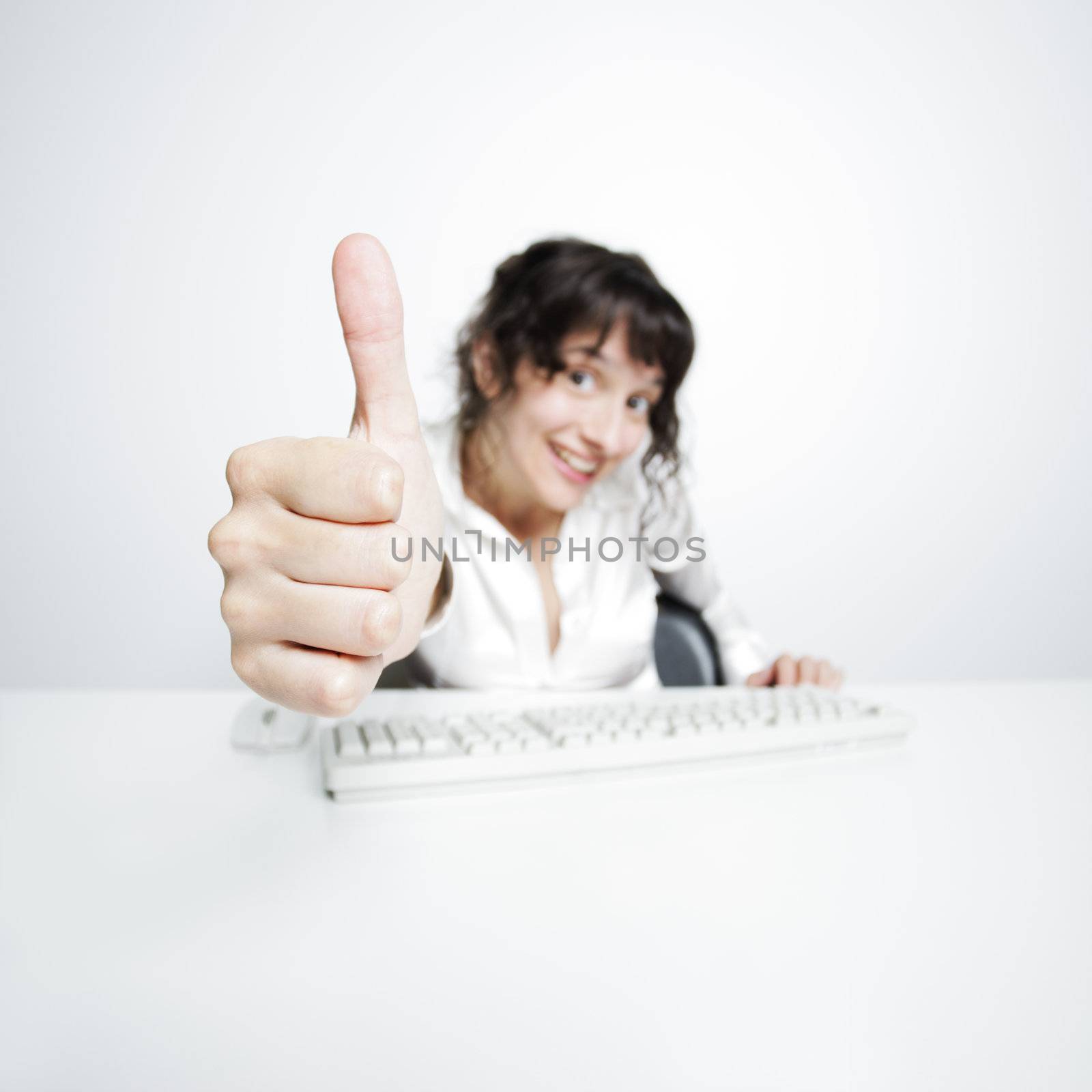 thumbs up shown by a happy, smiling young woman working at her office desk