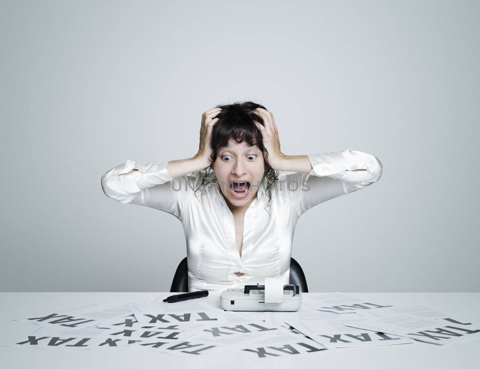 Young surprised woman at her desk frightened by taxes shouts out holding hands on  her head