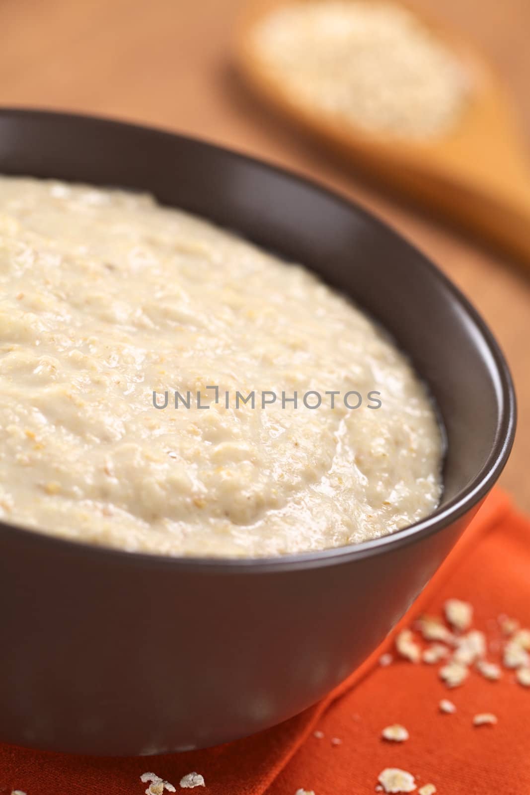 Bowl of cooked oatmeal porridge mixed with powdered maca or Peruvian ginseng (lat. Lepidium meyenii) with rolled oats on wooden spoon in the back (Selective Focus, Focus in the middle of the oatmeal porridge) 