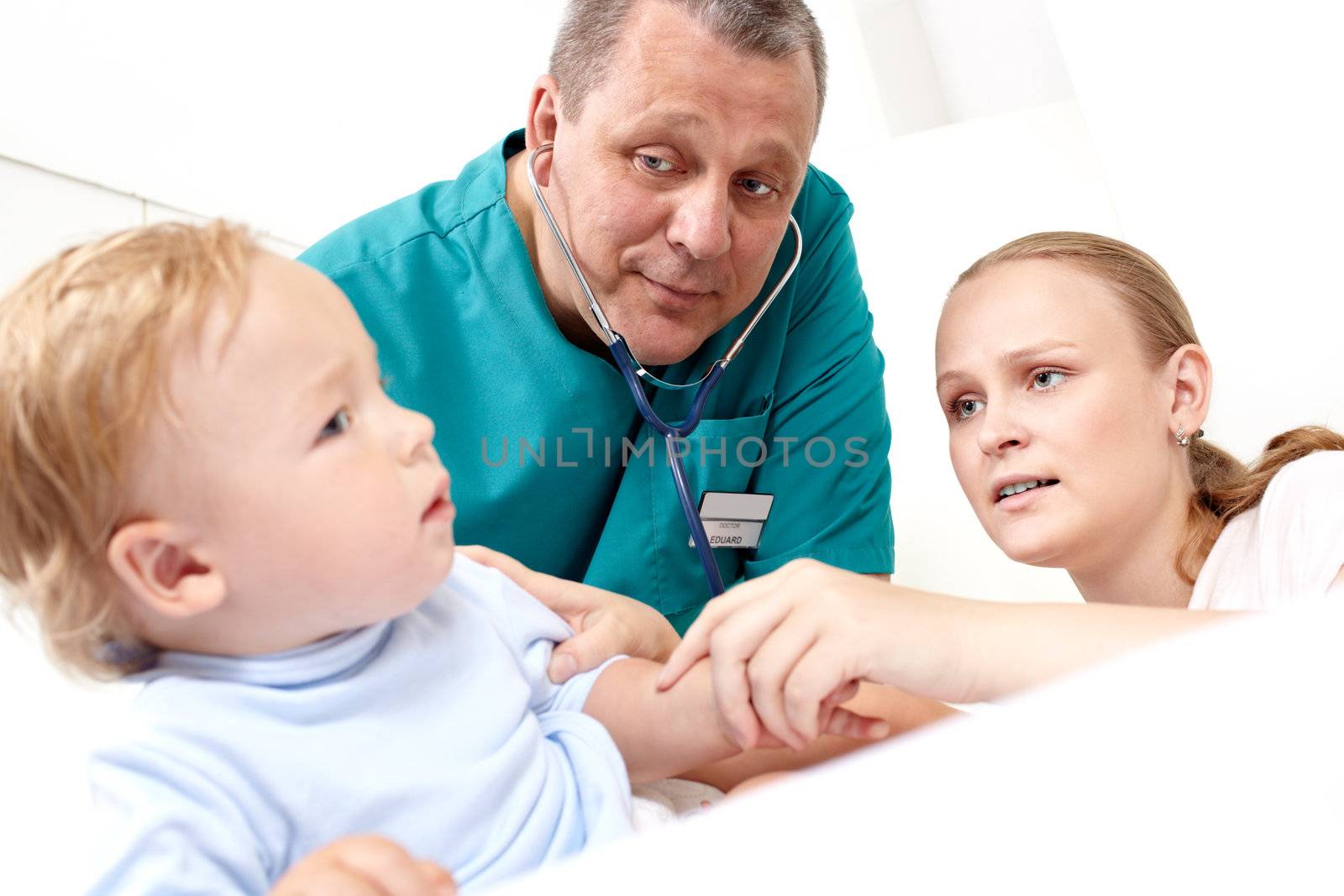 A middle-aged doctor is listening to a baby’s back with a stethoscope in  a medical study. The baby is 16 months old. The young mother is looking at her son with alarm. Caucasian. Focus on the mother and on the doctor.