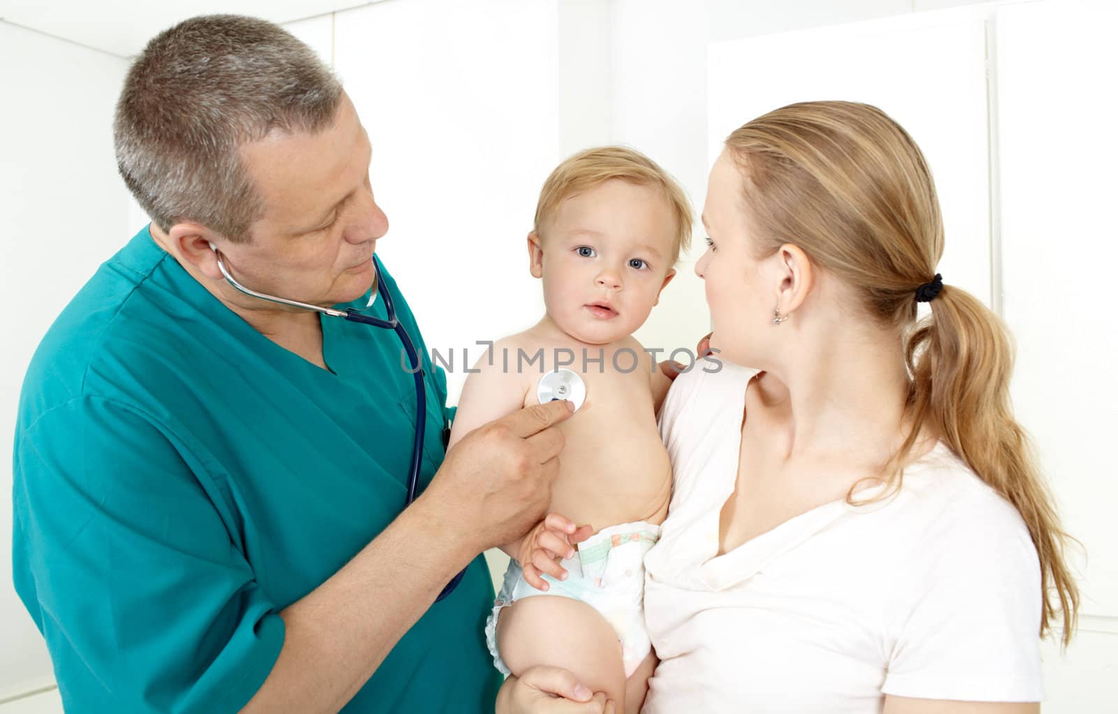 Doctor, middle–aged, in a green smock, is checking a baby with a stethoscope in a light medical study. The baby is 16 months old. He’s sitting on his mum’s laps. Caucasian, white.