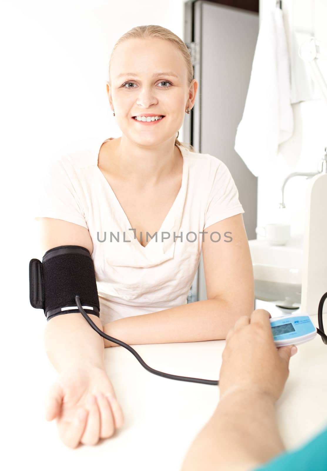 A young pretty girl of 25-30 in a white blouse is checking her blood pressure in a light medical study. She’s smiling and looking at the camera.